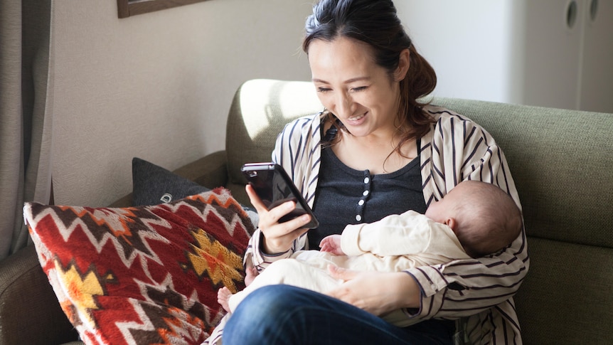 An asian woman sits on a lounge holding her sleeping baby and laughing at something on her phone