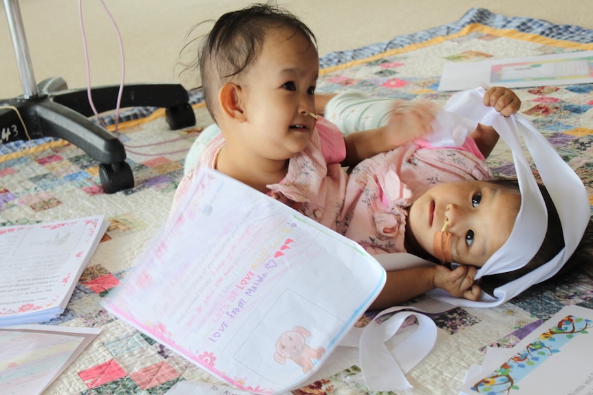 Conjoined twin girls lay on a blanket on the floor playing with paper