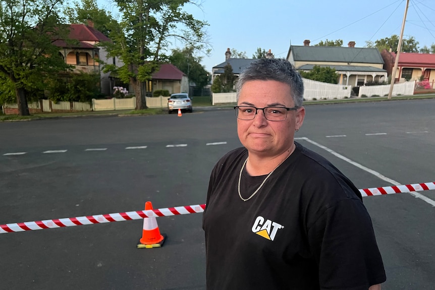 A bespectacled woman with short hair stands on a road that has been cordoned off with police tape.