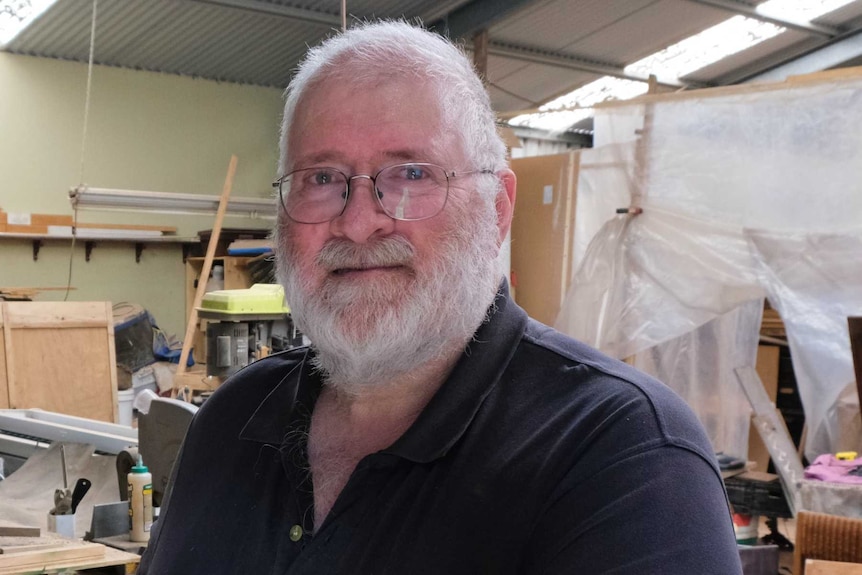 A man with white hair, a beard and glasses smiles standing in a wood working shop.