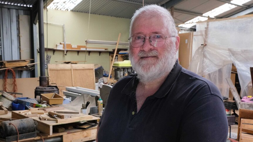 A man with white hair, a beard and glasses smiles standing in a wood working shop.