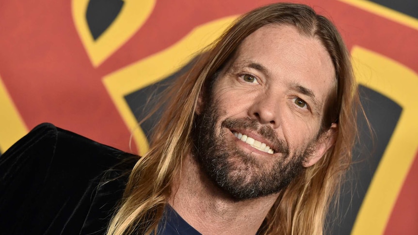 close up photo of taylor hawkins. he has a short beard, long hair and is smiling.
