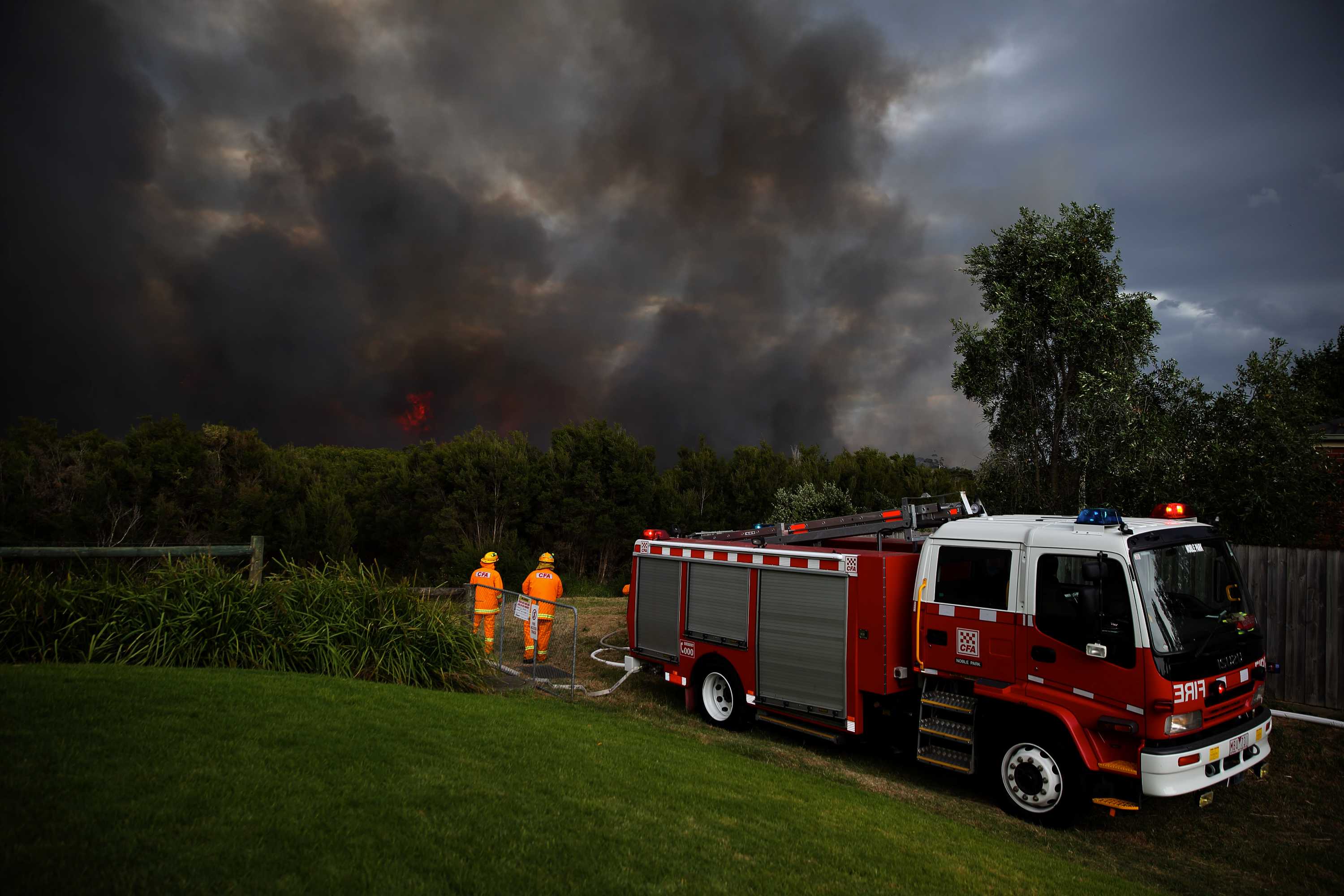 Two fire fighters stand next to a firetruck in front of a green lawn with a fire burning in the background