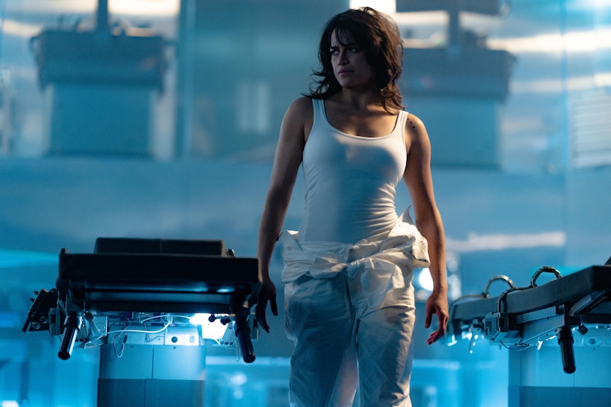 Michelle Rodriguez, a fit, brunette Latina woman wearing a white singlet and trackpants, walks through a blue-lit laboratory.