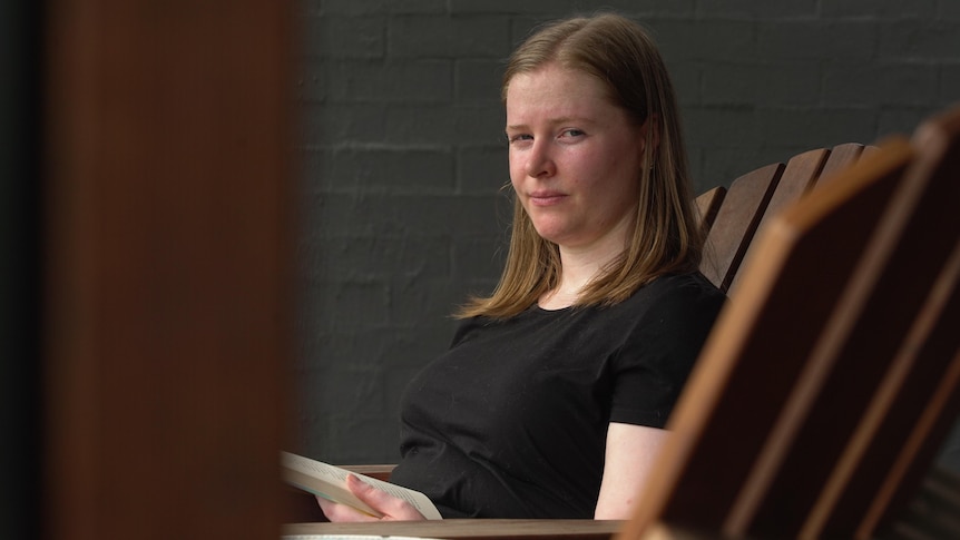 A woman sits on a deck chair, looking up from her book at the camera.