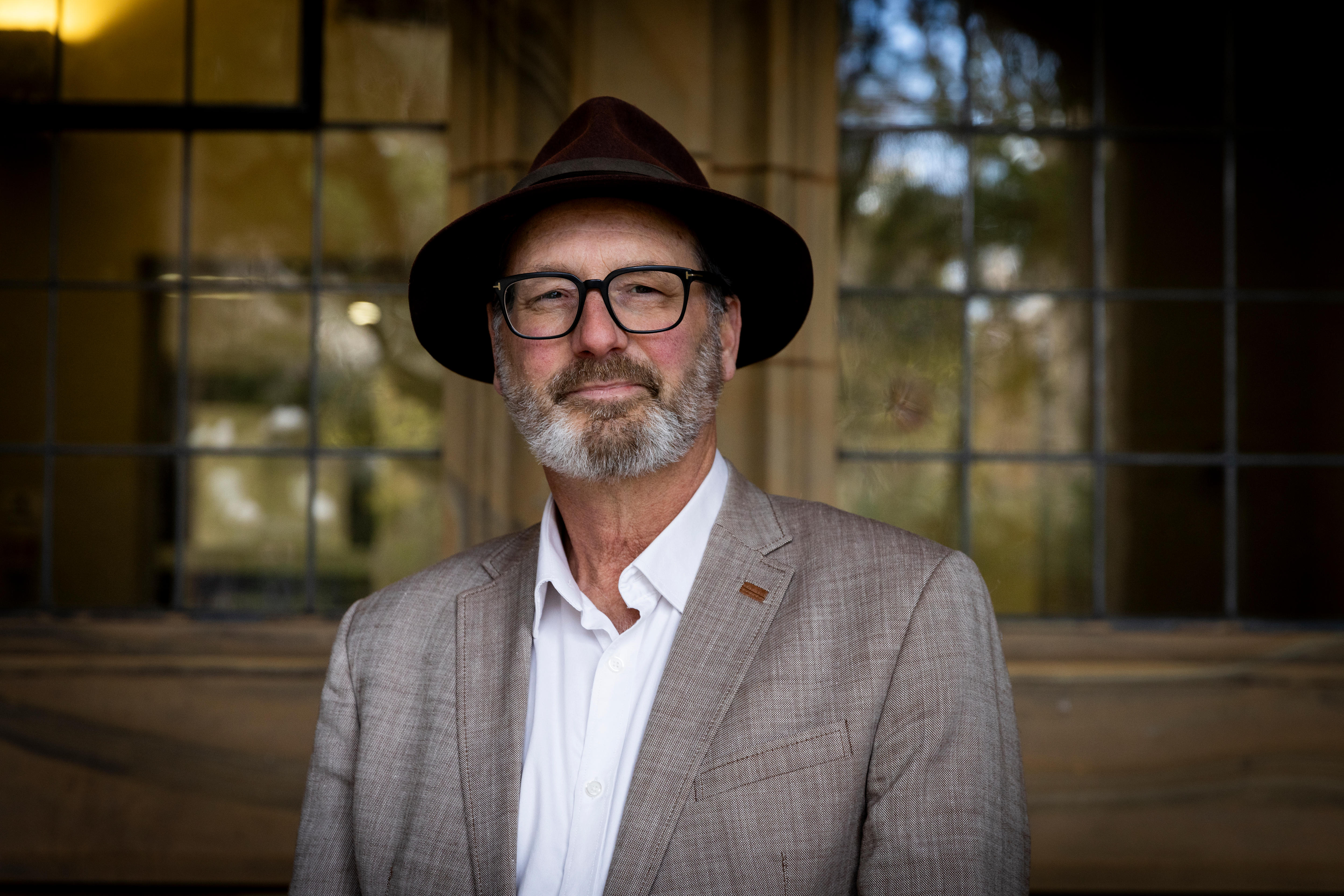 Grant Blashki smiles, standing in a suit, hat and glasses at a university campus.