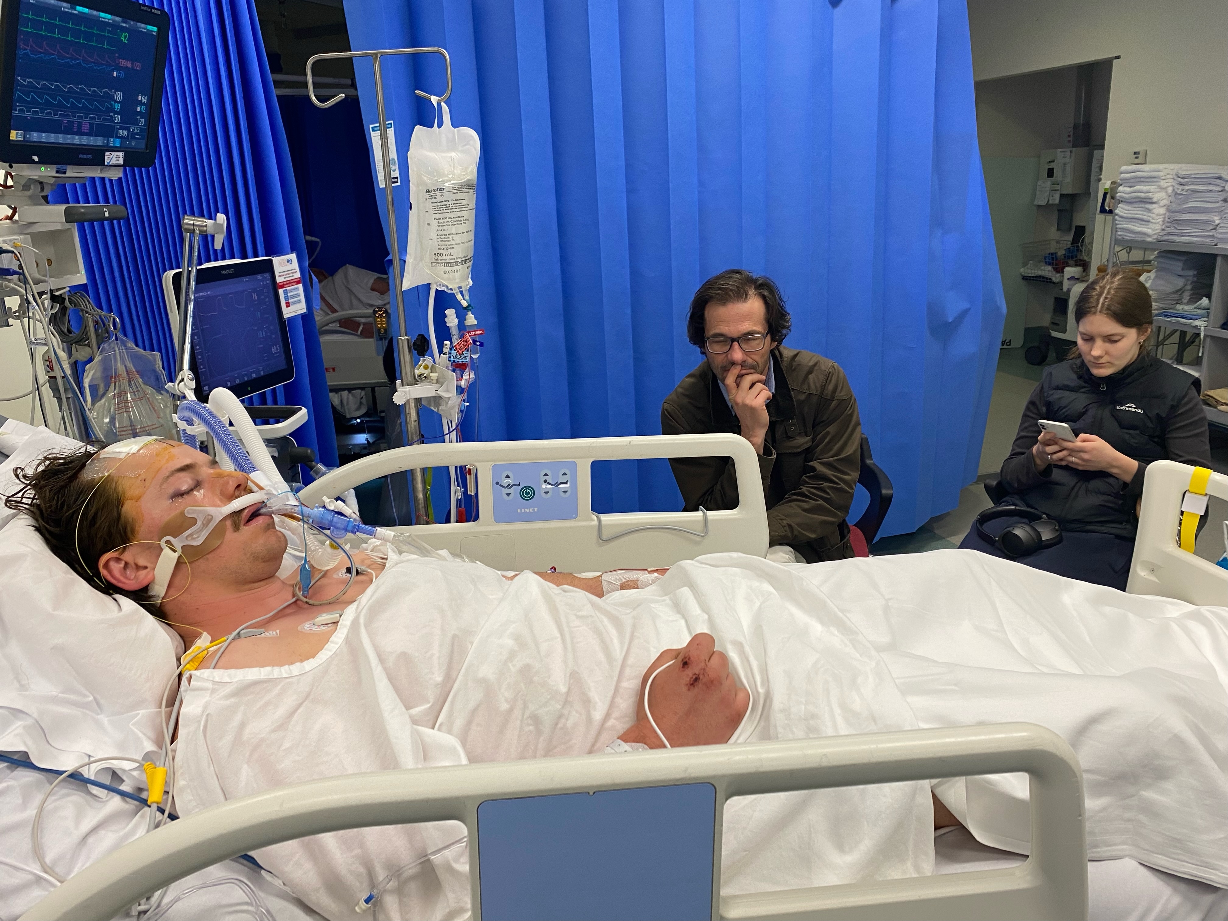 Xavier Sedger yace en una cama de hospital con tubos en la nariz. Lleva una bata de hospital.