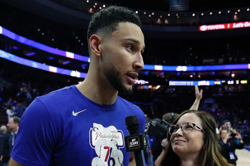 A basketballer stands at courtside talking with a reporter with a microphone in her hand.