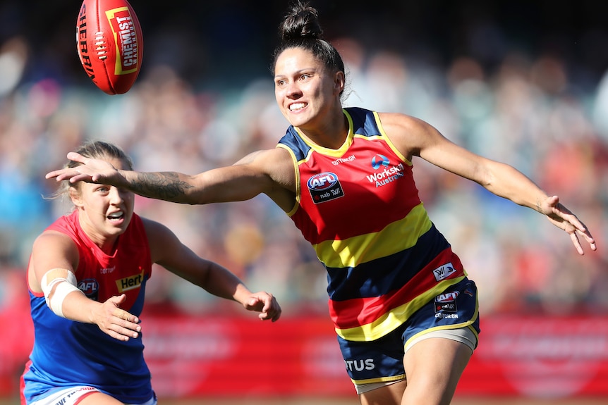 An AFLW player reaches out with her right hand extended to try to grab the football as a defender closes in. 