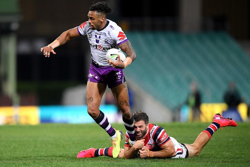 A Melbourne Storm NRL player holding the ball attempts to break a tackle of a Sydney Roosters opponent.