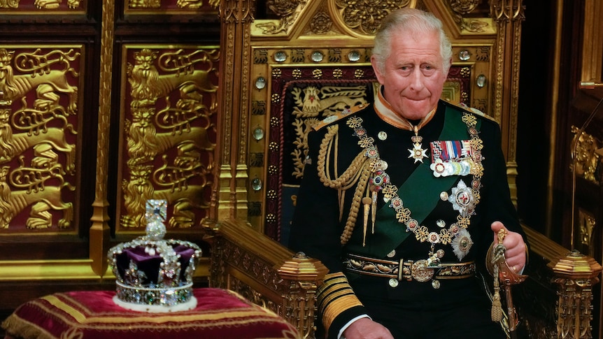 King Charles in full regalia seated next to the Queen's crown