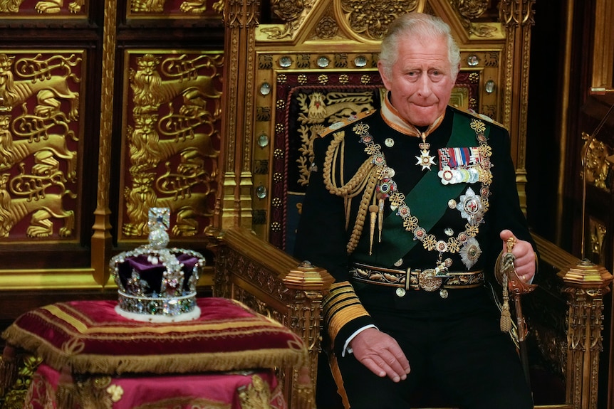 King Charles in full regalia seated next to the Queen's crown