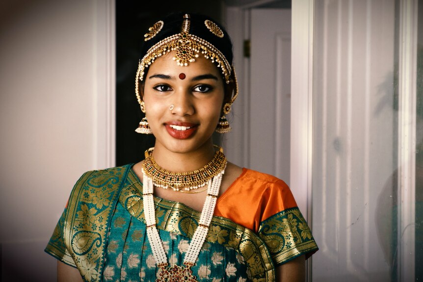 A young brown girl wearing a green and orange sari with beaded jewellery smiles confidently.