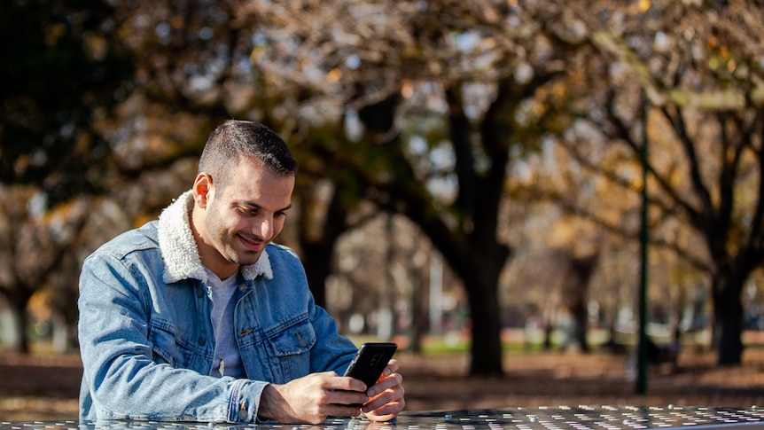 You view a man of Middle Eastern descent look into a phone while seated on a stainless steel bench in a park pictured in autumn.