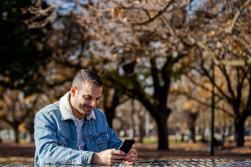 You view a man of Middle Eastern descent look into a phone while seated on a stainless steel bench in a park pictured in autumn.