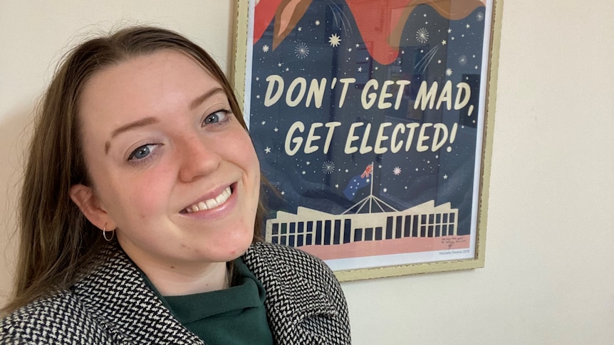 A young woman standing in front of an election poster that says "don't get mad, get elected"
