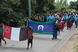 Parade on the street led by children and elders holding flags.