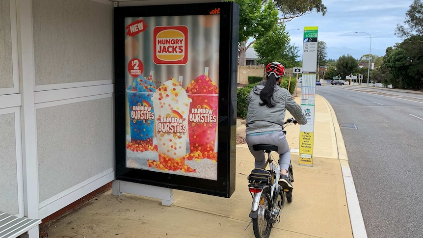 An advertisement for Hungry Jacks drinks at a bus stop, with a woman riding her bike off to the right.