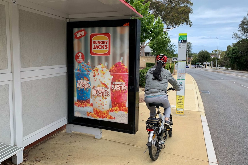 An advertisement for Hungry Jacks drinks at a bus stop, with a woman riding a bike off to the right.