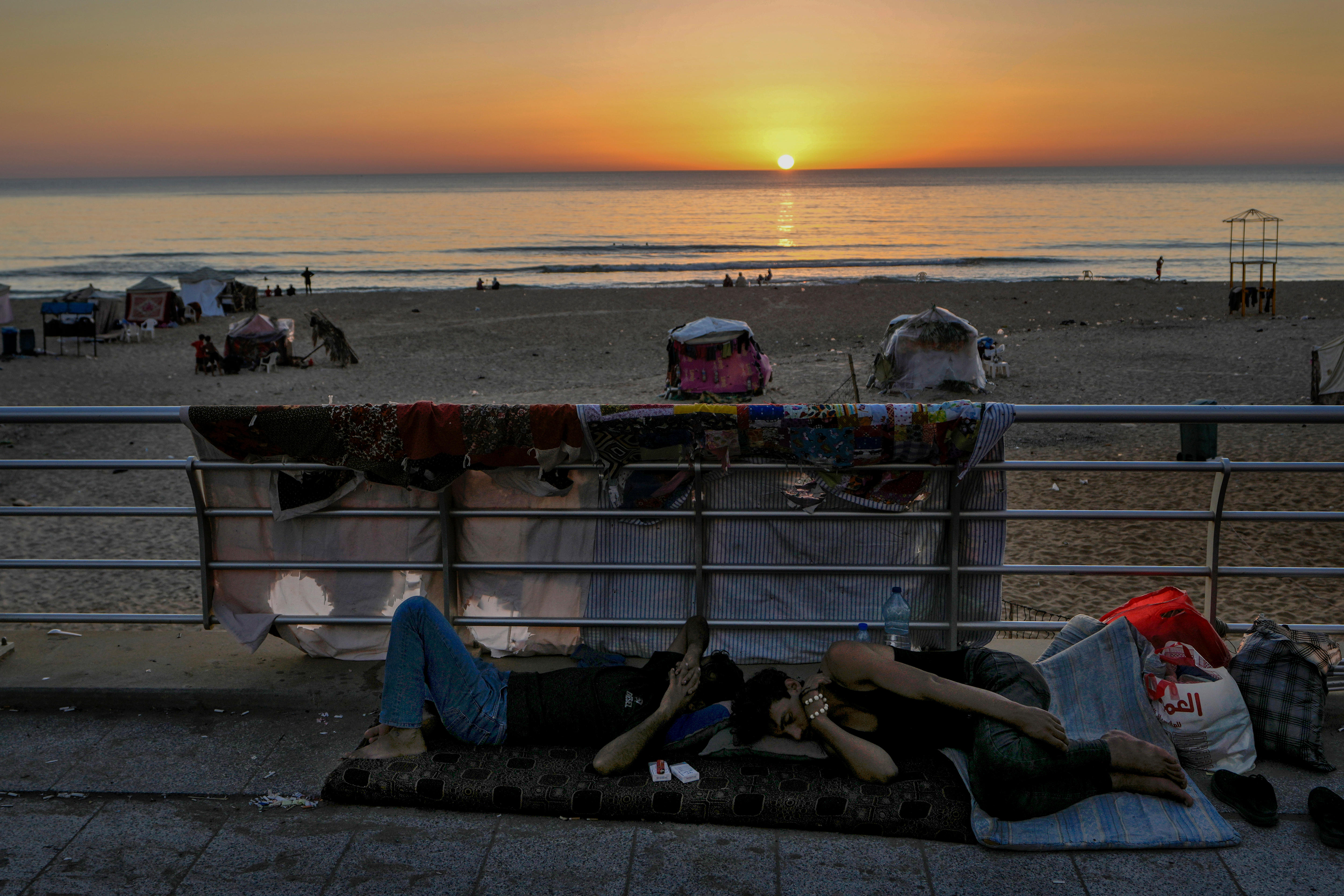 Two men sleep on thin mattresses next to a railing at a beach with the sun setting in the background