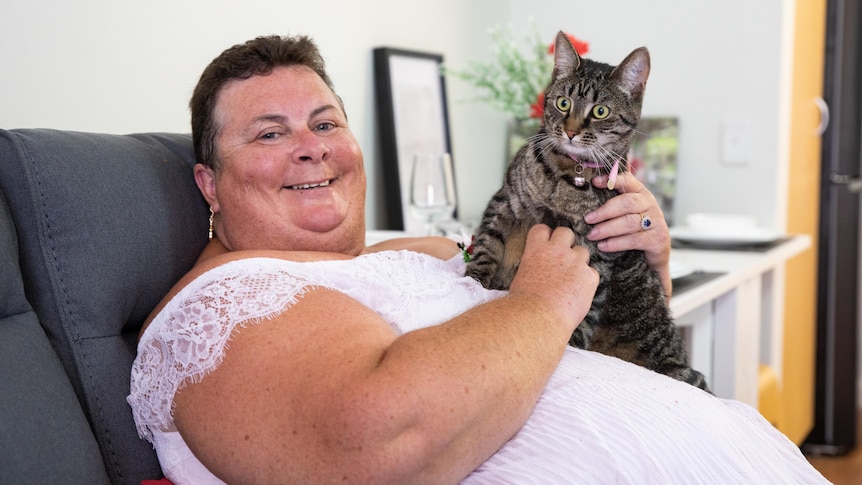 A woman sits smiling, stroking her cat. 