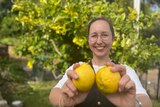 A woman stands smiling while holding two lemons in the foreground. 