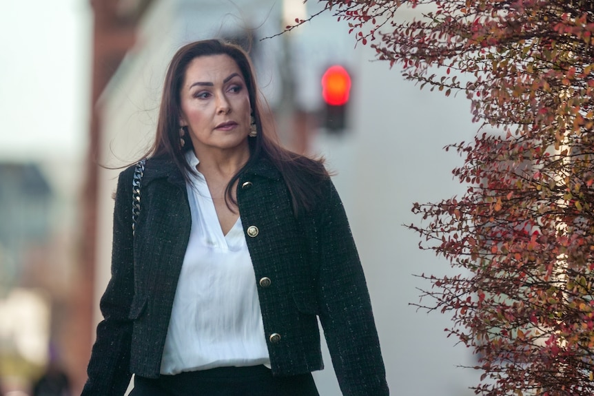 A woman with windswept, dark hair wearing a black coat and white blouse turns a street corner with autumn foliage.