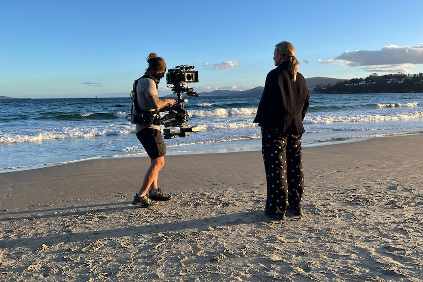 Marta Dusseldorp, a blonde 51-something woman, stands on a beach, facing away, as a cameraman films her.