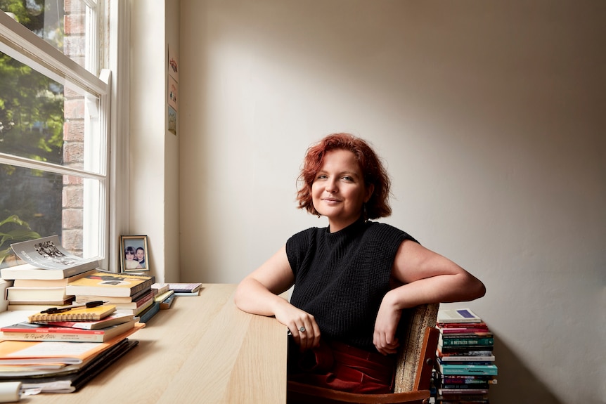 A white woman with chin-length wavy red hair sits at a desk by a window with books stacked in piles on top