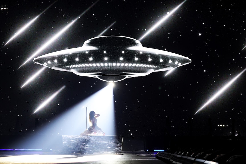Taylor Swift sings kneeling on a platform with the spaceship floating above