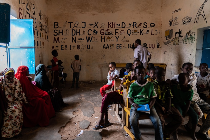 Children sit in a classroom with a dusty floor, and the alphabet painted onto the walls. One child looks directly to camera