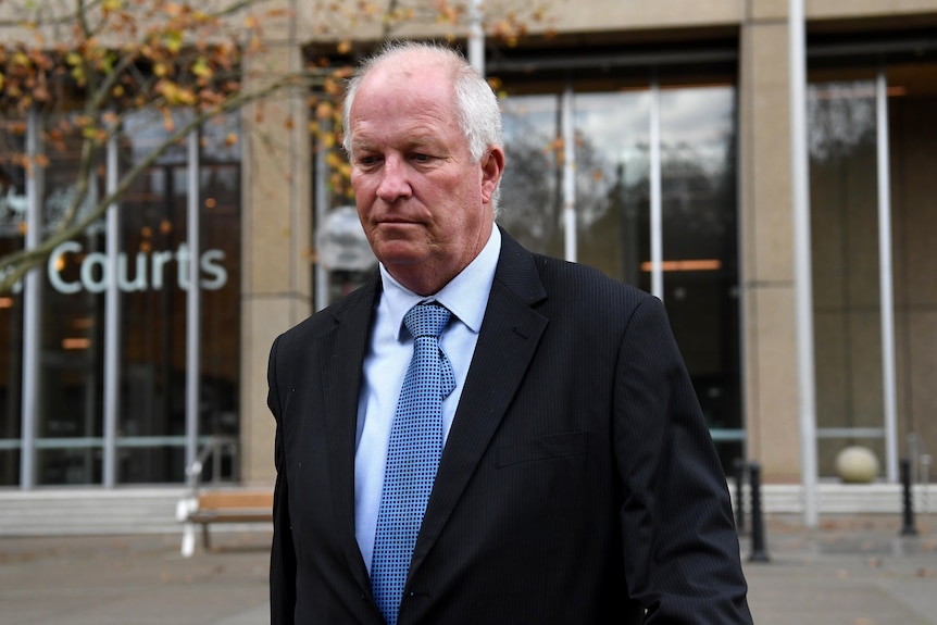 Man wearing black suit jacket and blue tie looks down