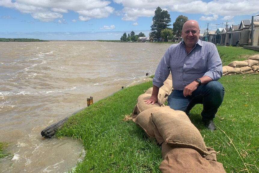 A bald man squatting next to sandbags and a lake