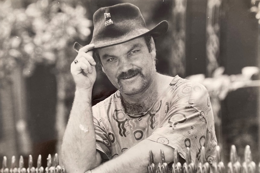 A man wearing a t-shirt and wide brim hat leans over a fence posing for a photo