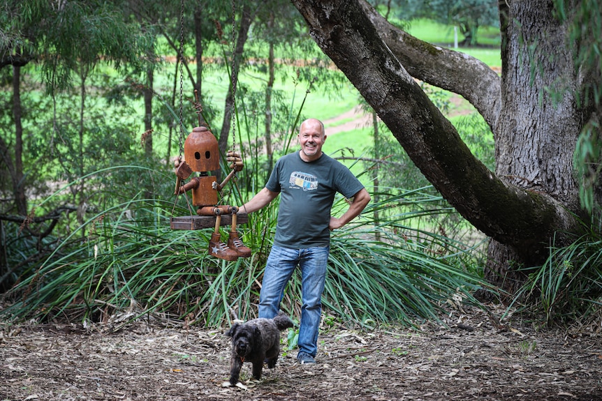 A man poses next to a metal robot on a swing, with his dog at his feet.