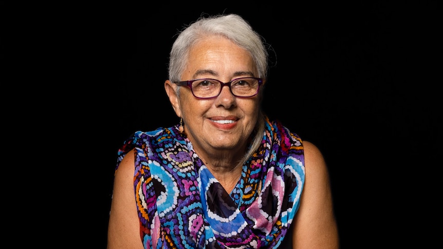 Portrait of Sue Pinckham, wearing a blue/purple dress, looking at the camera against a black background.