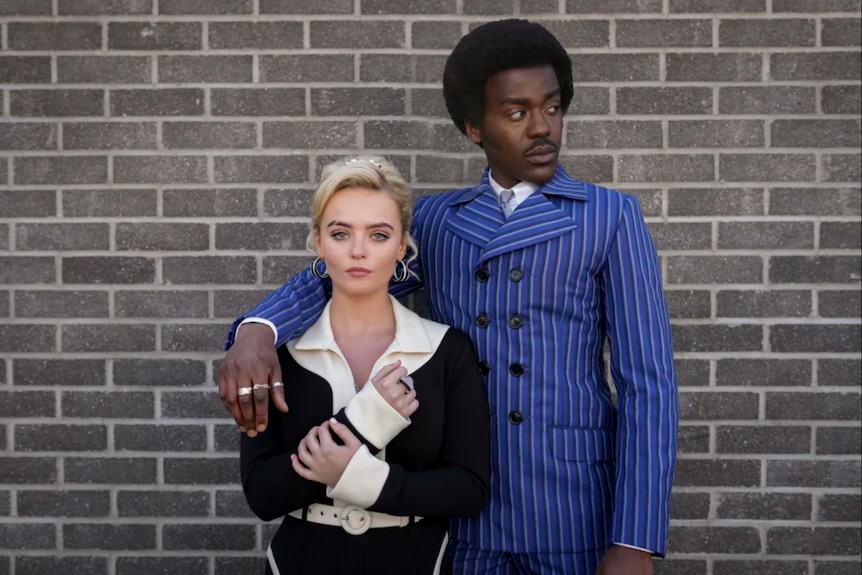 Millie Gibson and Ncuti Gatwa stand together in 70s style outfits in front of a brick wall