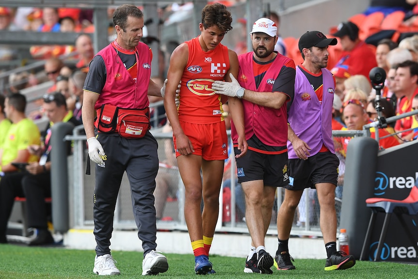 A concussed Gold Coast Suns AFL player walks off the field while being helped by two trainers.