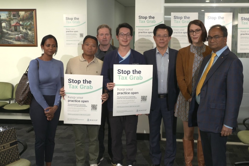 A group of people stand in a GP clinic waiting room with some holding signs saying "stop the tax grab".