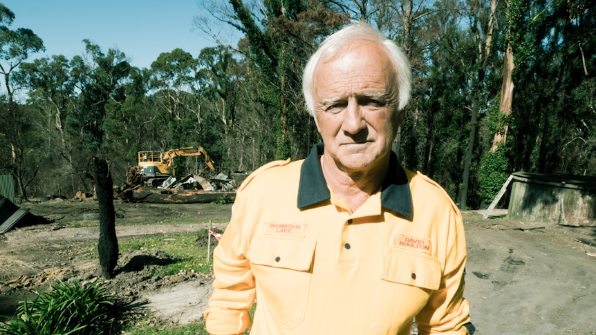 Man in RFS uniform standing at his property with an excavator removing fire debris in the background.