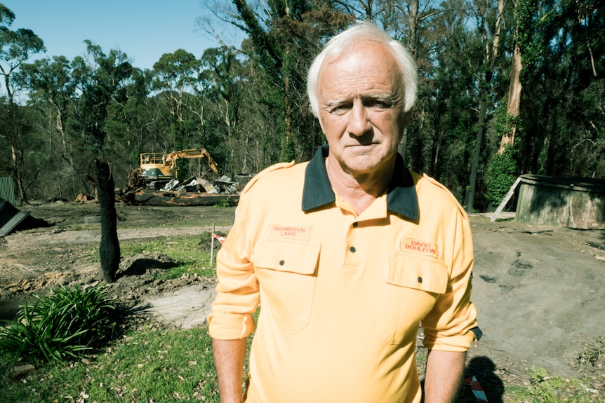 Man in RFS uniform standing at his property with an excavator removing fire debris in the background.