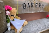 A teddy bear is placed on a wooden seat outside a bakery