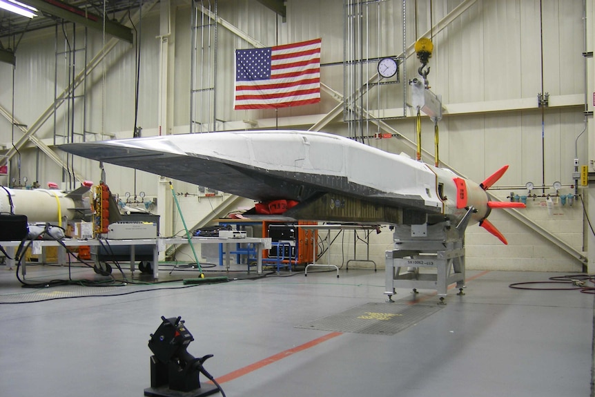 In an air hangar, an aircraft shaped like a wedge with no windows sits on a platform with a large US flag on the wall behind it.