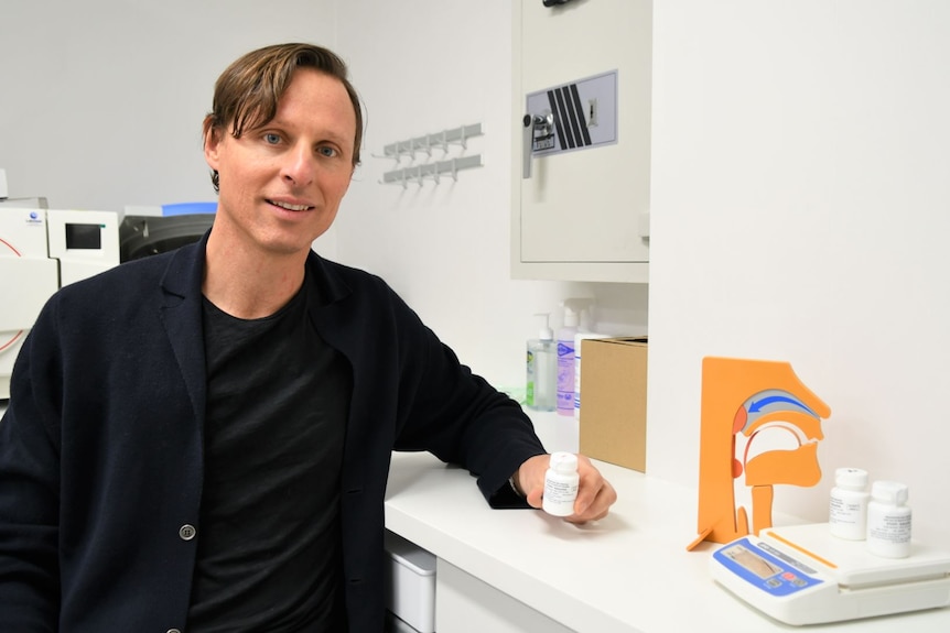 A male professor with a side part wearing black leans over a counter holding a white pill bottle