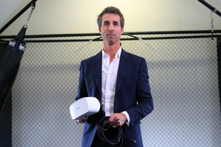 A man in a blue suit, holding a virtual reality headset, standing in front of an indoor chain link fence.