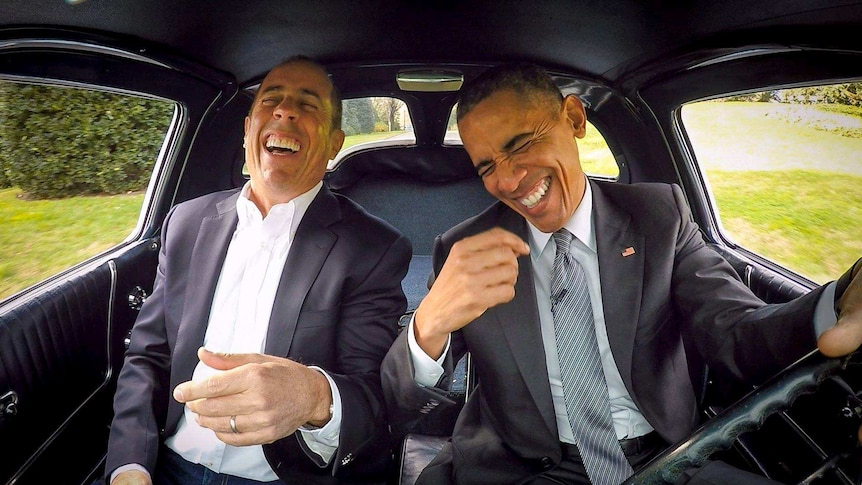 Barack Obama laughs while driving a 1963 Corvette with Jerry Seinfeld in the passenger seat.