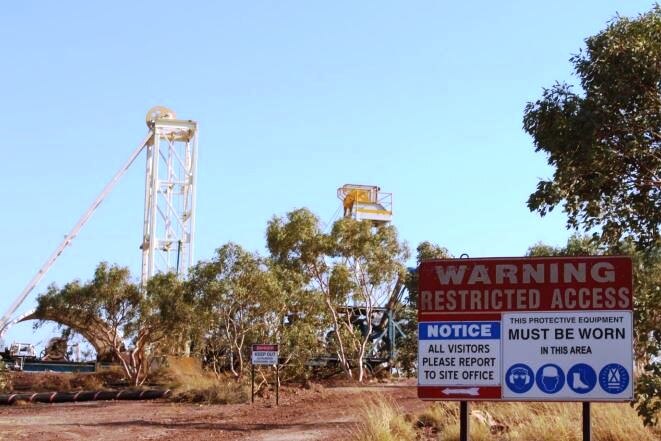 A shot of bushland with two drill rigs in the background. A safety sign is in the foreground.