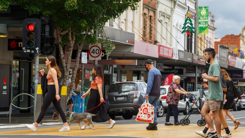 Pedestrians walk across a crossing on a Moonee Ponds street, with Christmas decorations hanging from shops.