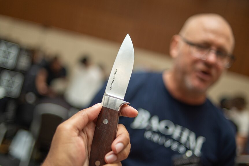 A short-bladed knife held in a hand with a man in the background.