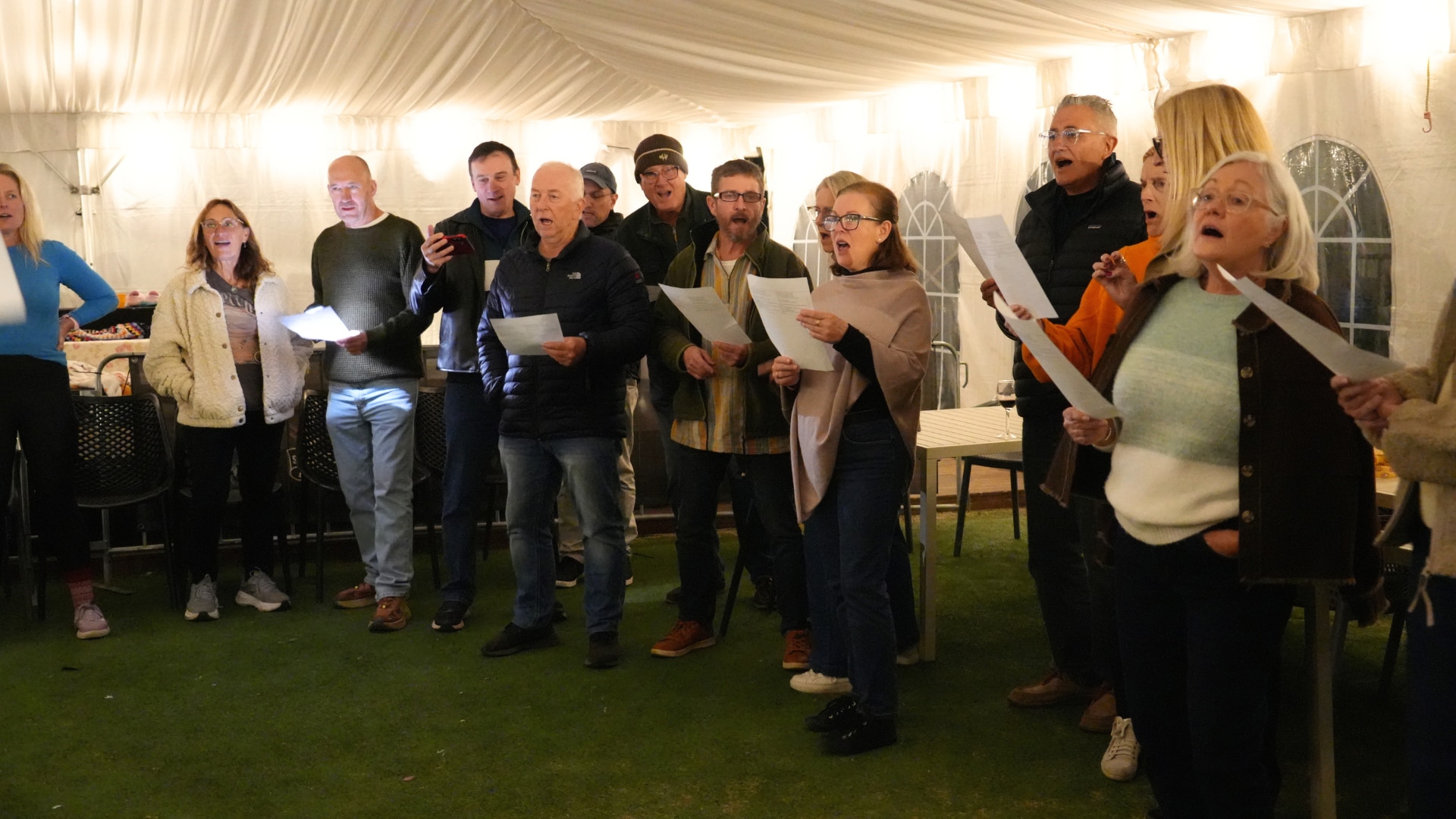 A group of men and women under a white marquee holding piece of paper with lyrics and singing.